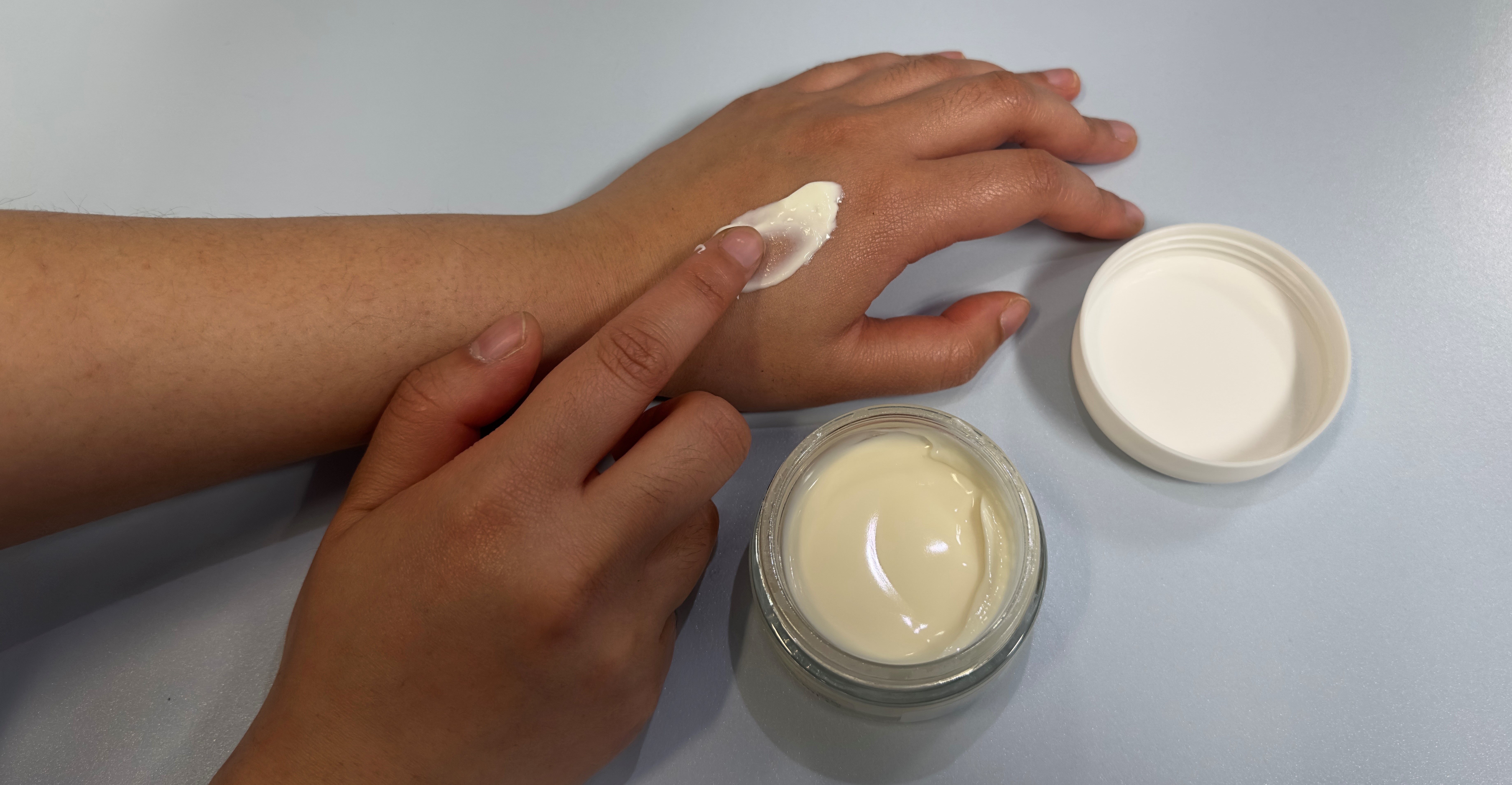 Das Bild zeigt eine Hand auf der mit der anderen Hand eine weiße Creme verteilt wird. Daneben steht das geöffnete Produkt.